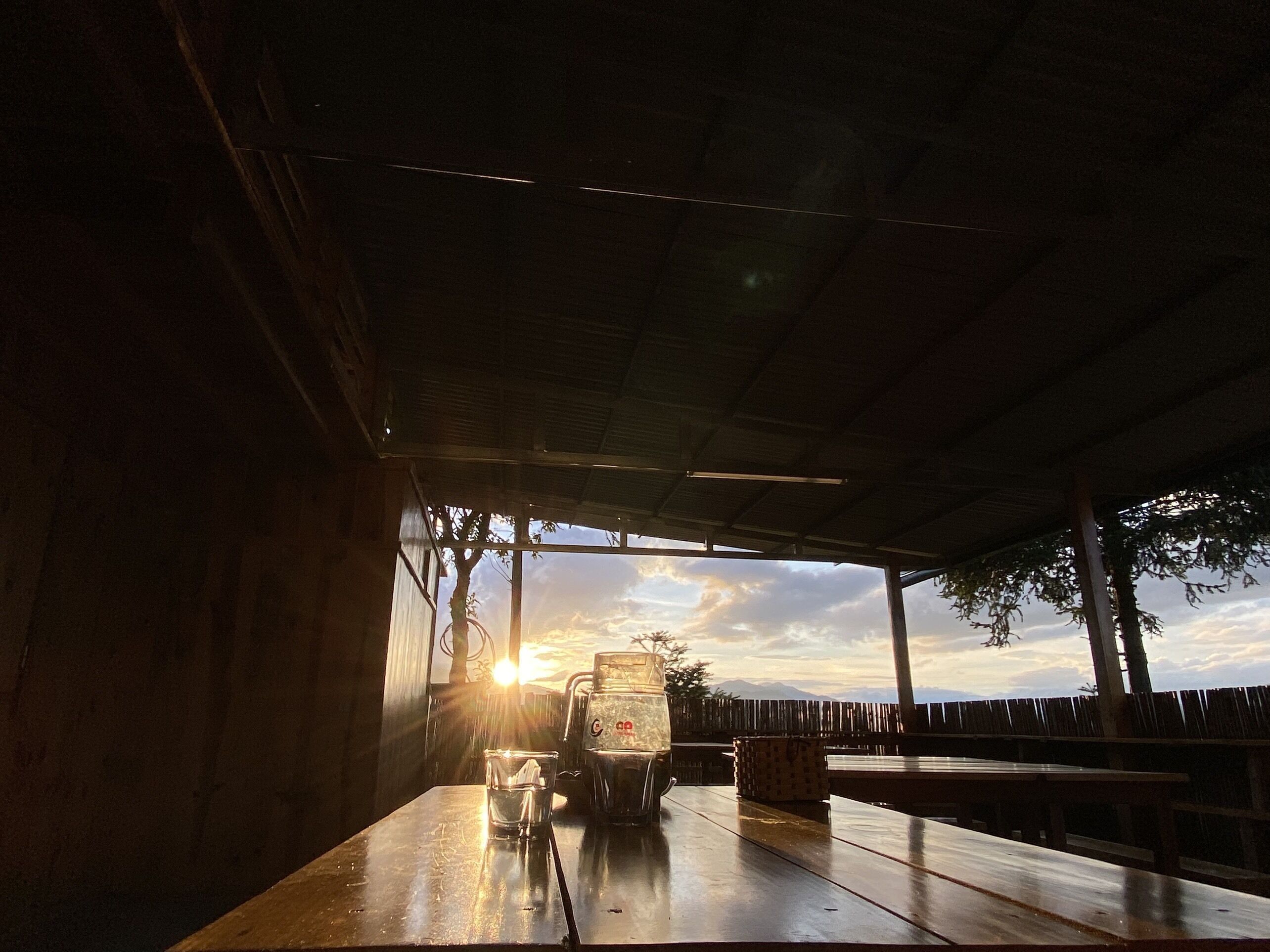 An outdoor seating area with a view of the sunset, a table with a glass of water, and a stack of plates.