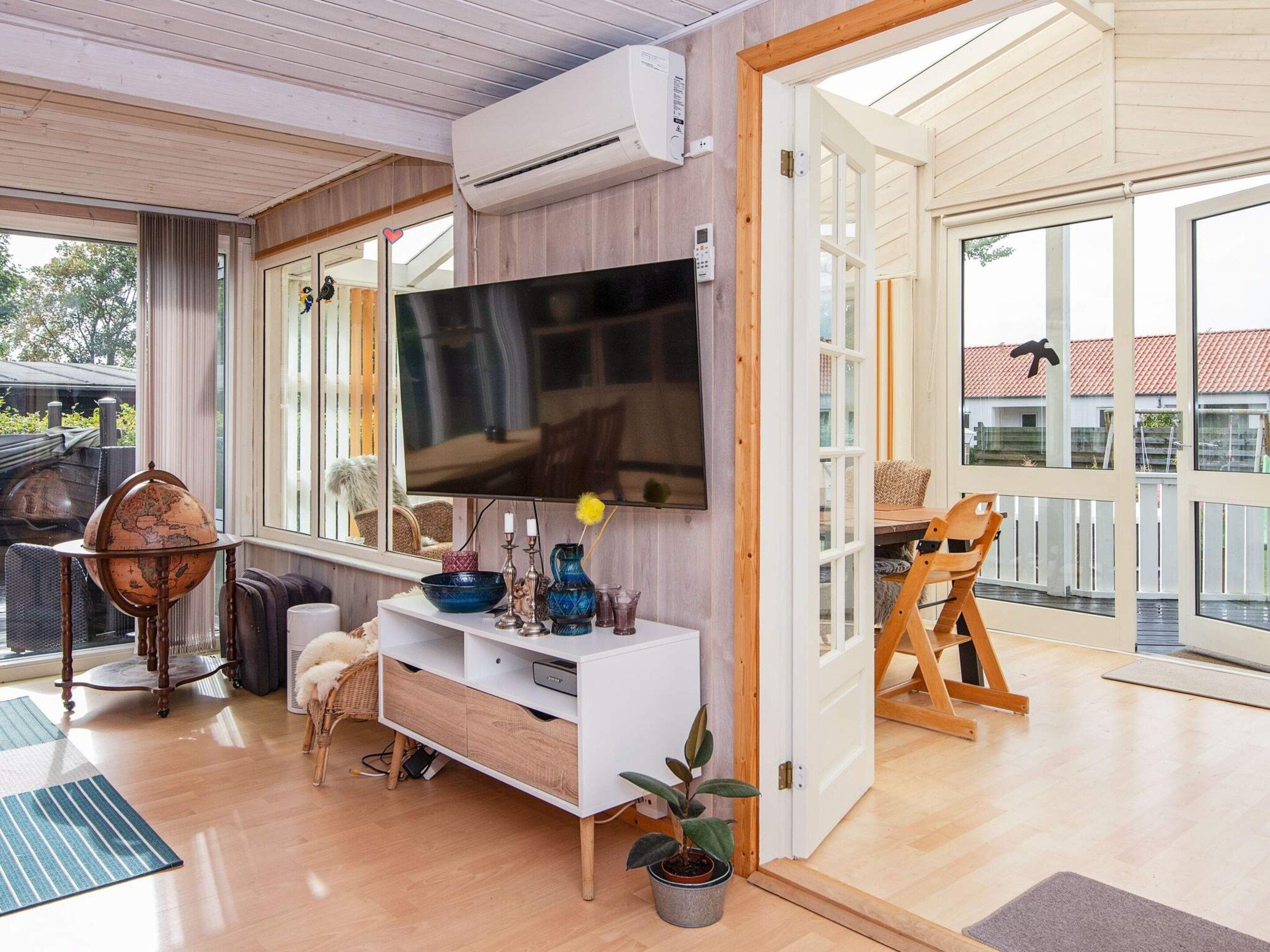 A modern living room with a large flat-screen TV, wooden flooring, a white entertainment unit, and a large window offering a view of the outdoors.