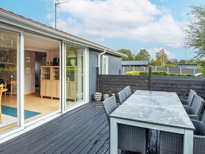 Una terraza con mesa y sillas, una puerta corrediza de vidrio que da a un espacio interior con cocina, y una valla de madera.
