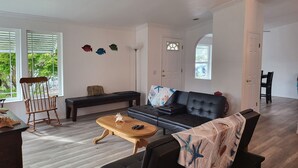 Een woonkamer met een bank, een houten salontafel, een schommelstoel en een bankje. Je ziet afbeeldingen van vissen op de muur, een deur en een raam met jaloezieën.