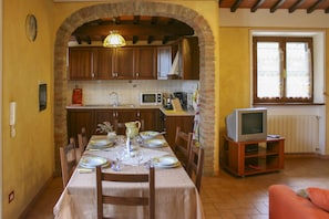 A dining area with a table set for four, a kitchen with wooden cabinets, a microwave, and a television on a stand.