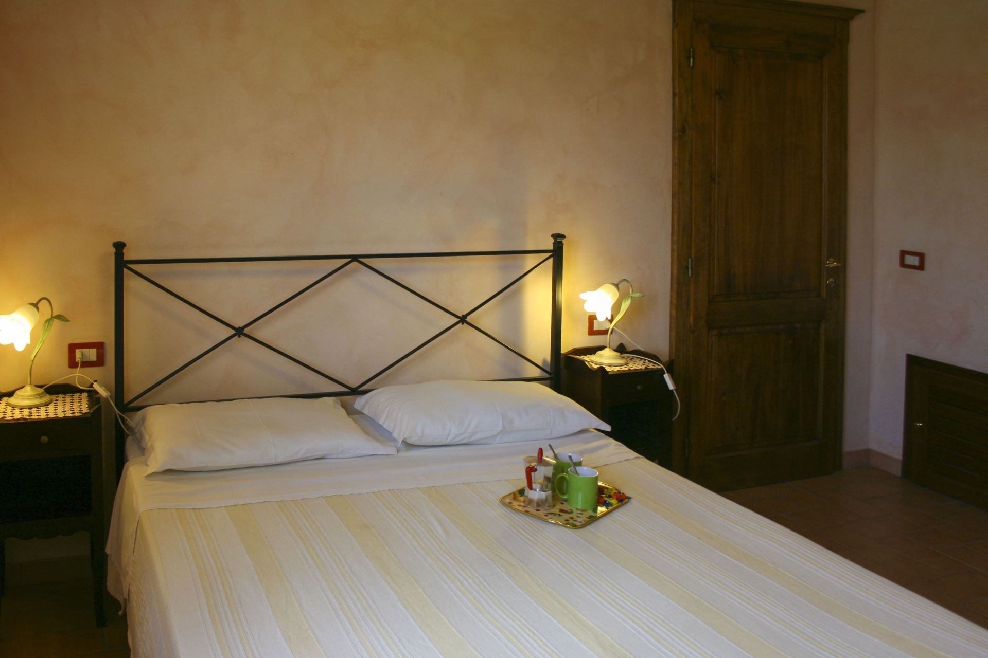 Ein ordentlich bezogenes Bett mit einer gestreiften Tagesdecke, zwei Nachttische mit Lampen, eine Holztür und ein kleines Tablett mit Tassen und einem Löffel auf dem Bett.