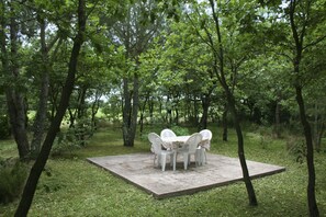 A square outdoor seating area with four white plastic chairs and a table, surrounded by trees in a natural setting.