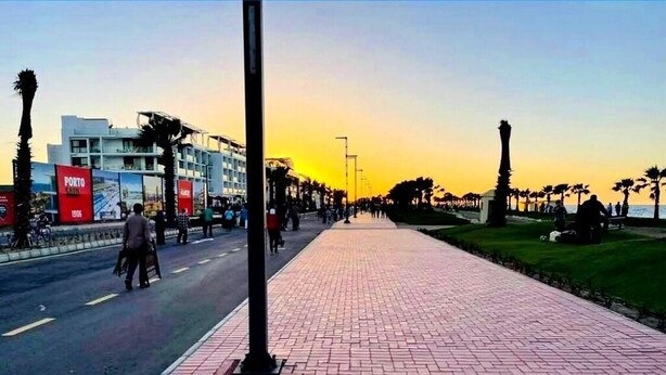A paved walkway with a brick pattern, lined with palm trees and a building with a red sign on the left. - Elegant Family Chalet with Pool View in Porto Said Resort (Port Said Governorate)