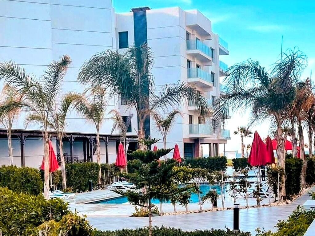 A modern hotel with a pool area, surrounded by palm trees and red umbrellas.