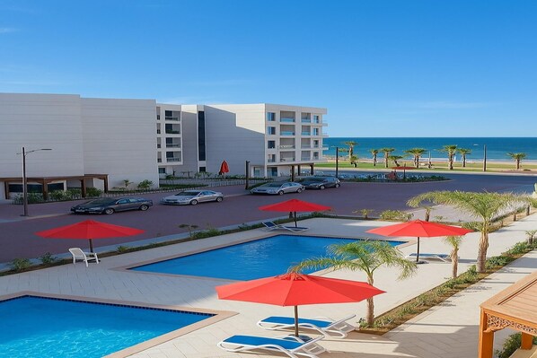 Zona de piscina de hotel con sombrillas rojas, sillas de descanso y vistas al océano.