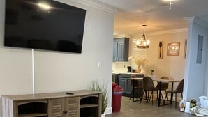 A living room with a large flat-screen TV mounted on the wall, a wooden entertainment unit, a beige sofa, and a dining area with a table and chairs.