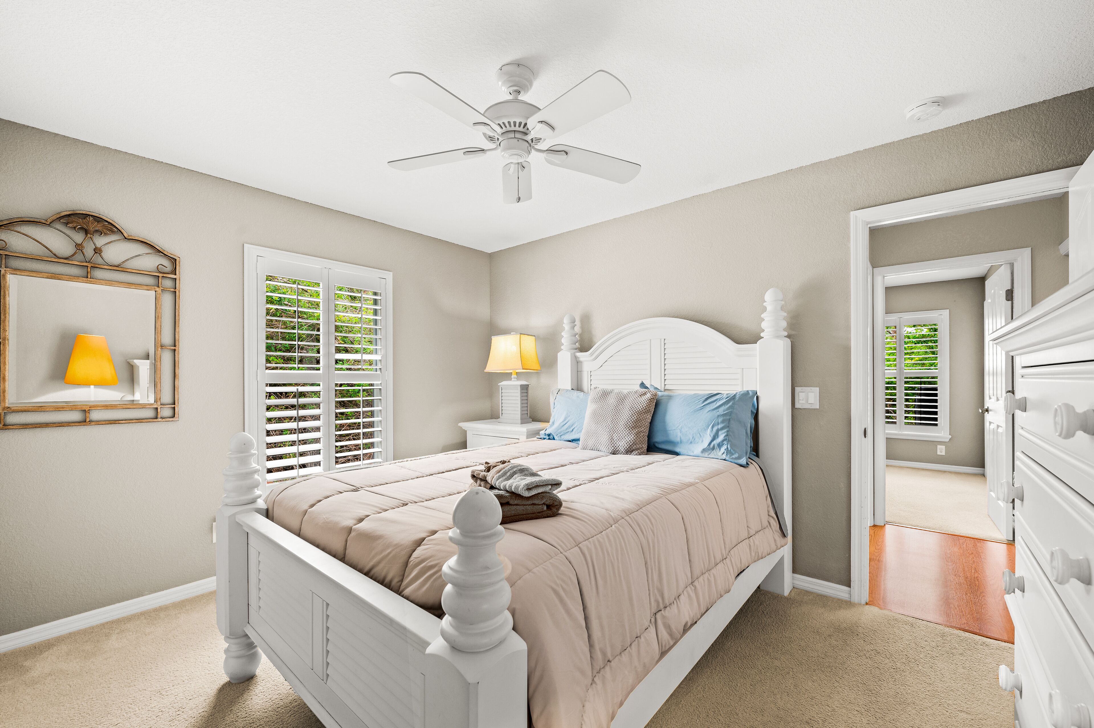Quarto com cama branca, paredes bege, ventilador de teto e janela com persianas.