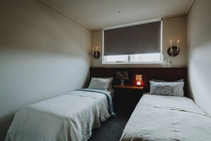 Une chambre avec deux lits simples, une fenêtre avec un store, une table de chevet avec une lampe et des appliques murales.