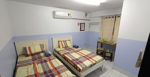 A room with two beds, a desk, a chair, an air conditioning unit, and a window with curtains.