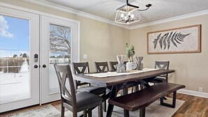 Un coin repas avec une table en bois, des chaises foncées et un banc. Un tableau représentant une fougère, encadré, est accroché au mur, et un luminaire est suspendu au plafond au-dessus de la table.