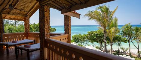 A covered outdoor area with a view of the ocean, featuring a wooden table and chairs, and a thatched roof.