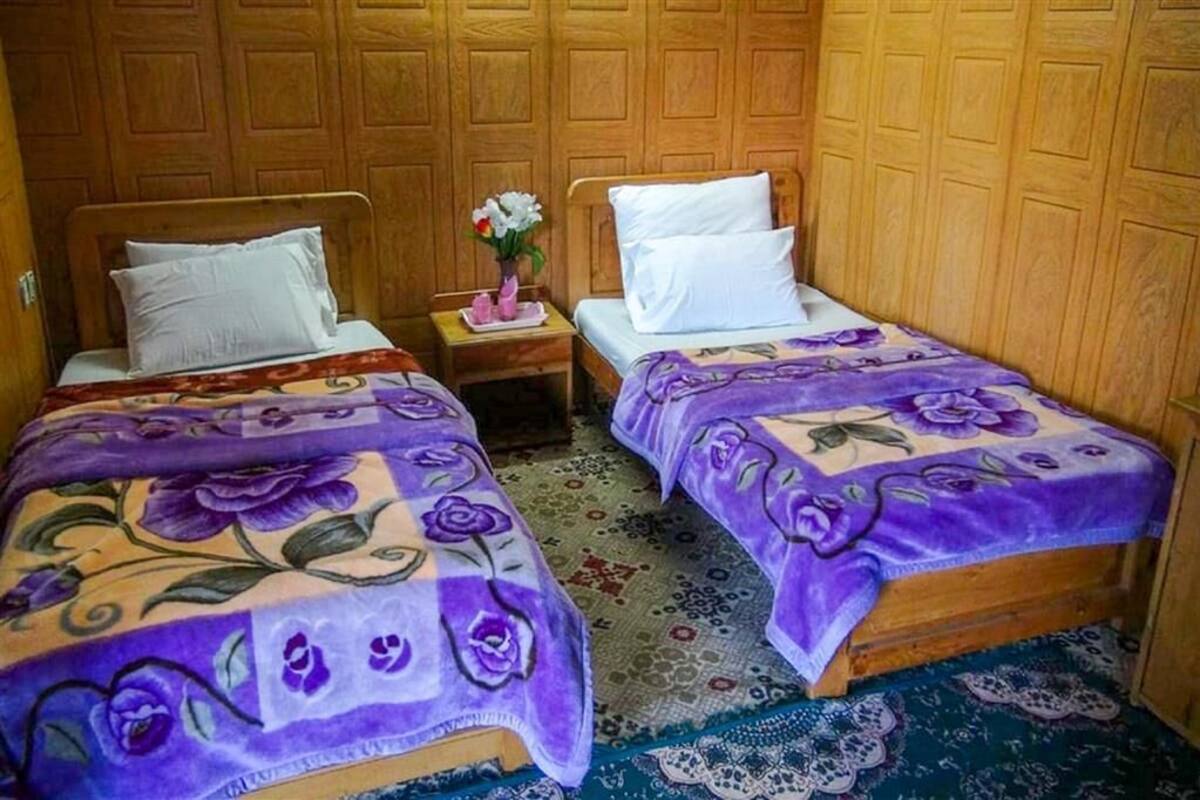 Una habitación con dos camas de madera, cada una con una colcha floral morada y almohadas blancas. Hay una mesita de noche con un florero de flores entre las camas.