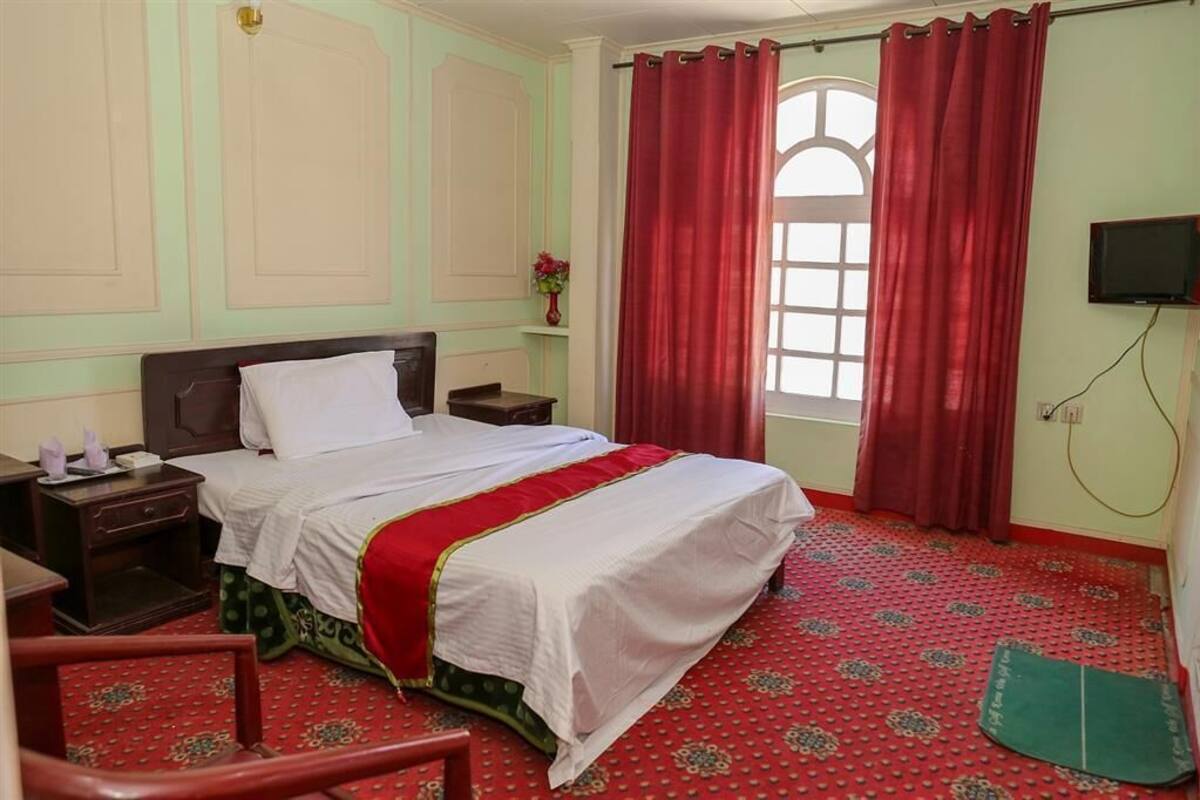 Una habitación de hotel con cama, mesitas de noche, televisor y ventana con cortinas rojas.