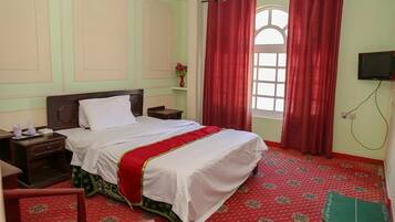Quarto de hotel com cama, mesas de cabeceira, televisão e janela com cortinas vermelhas.