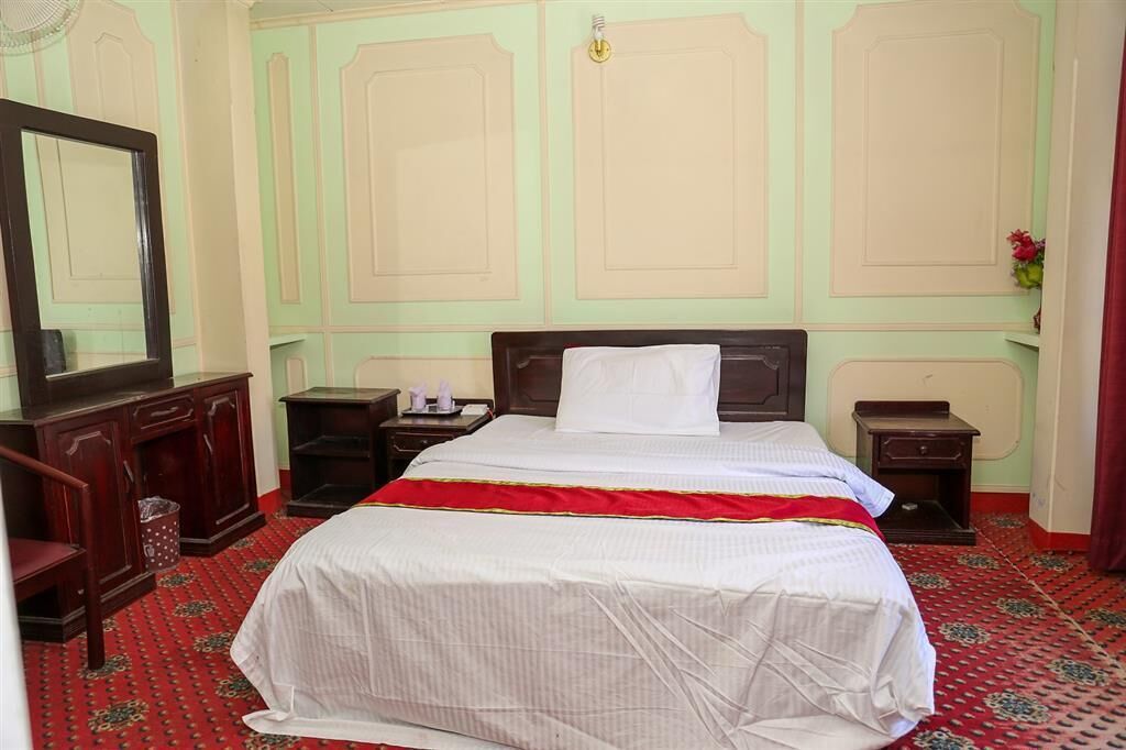 Ein Hotelzimmer mit einem Bett, einem hölzernen Schminktisch, einem Spiegel und einem roten Teppich mit Muster.