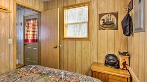 A room with wooden walls, a bed with a patterned bedspread, a window with blinds, a wall-mounted fan, and a door with a red and yellow curtain. - Secluded Remer Cabin w/ Lake Access! (Remer)