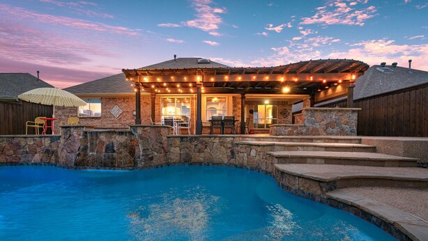 A backyard with a swimming pool, stone steps, and a covered patio area with outdoor furniture. - Unique heated pool & spa outdoor kitchen, game room near DFW  sleeps 10-12 (Lewisville)