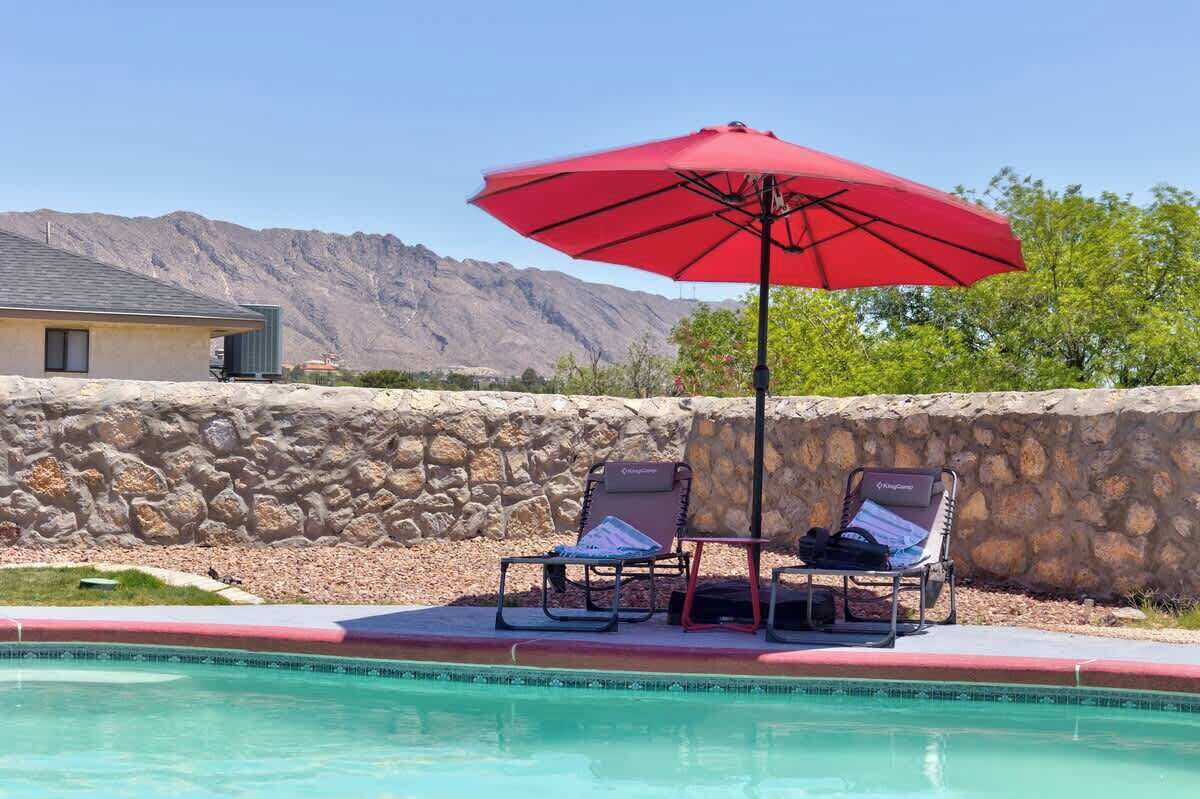 A pool area with two lounge chairs and a red umbrella, set against a stone wall and mountains in the background.