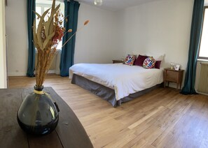 A bedroom with a wooden floor, a bed with white bedding and colorful pillows, a bedside table with a lamp, and a vase with dried flowers on a wooden table.