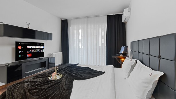 A modern hotel room with a bed, a TV on a stand, a bedside table, and a window with sheer curtains. - Toia Studio (Brasov)
