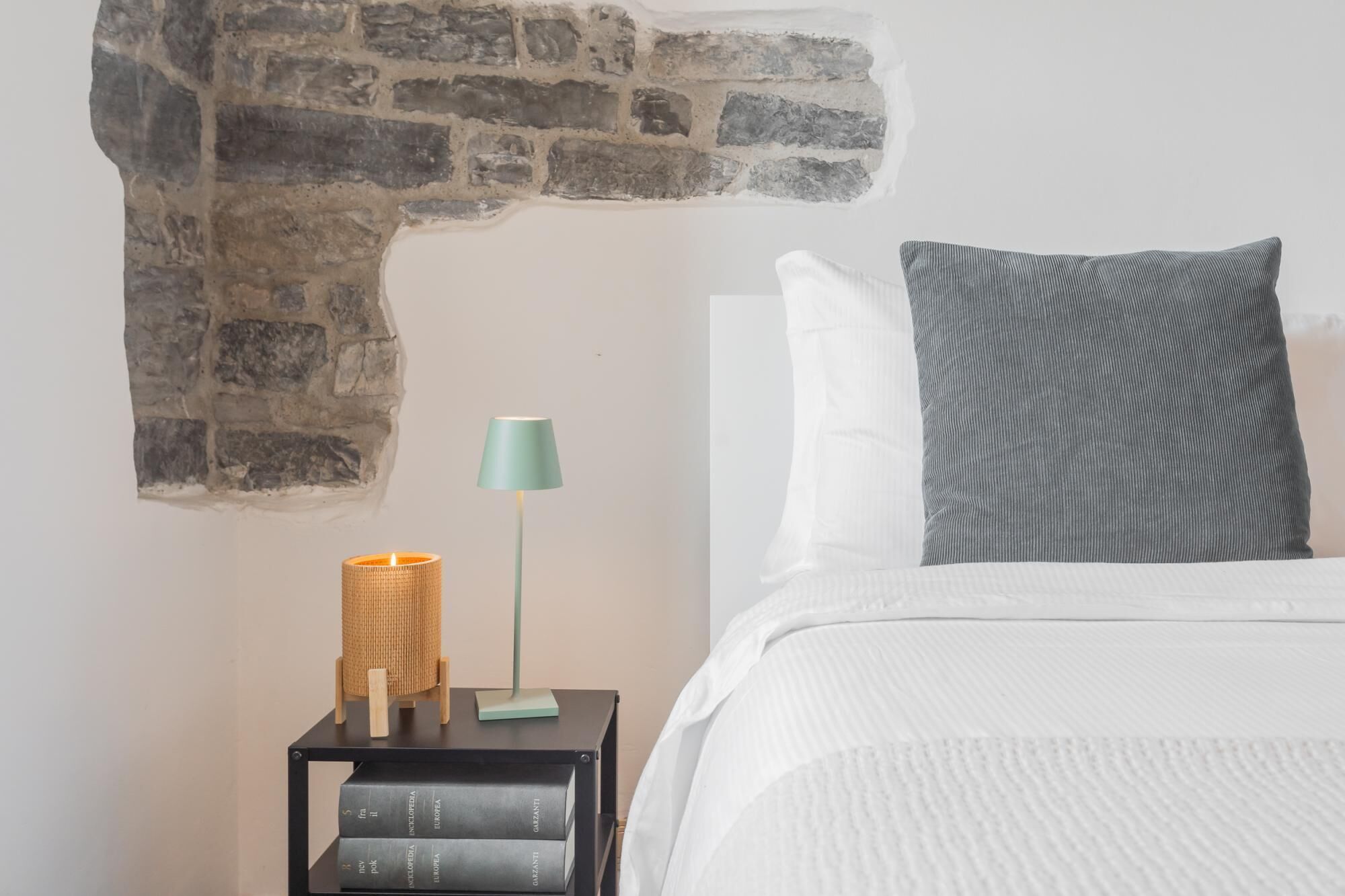 A neatly made bed with white linens and a grey pillow, a small black nightstand with a lamp and books, and a stone wall feature.