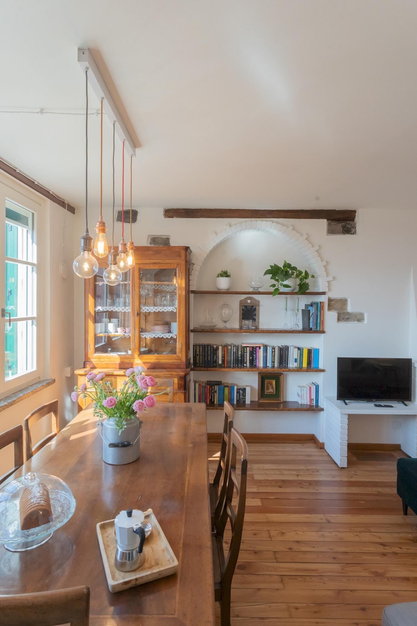 A dining area with a wooden table, chairs, a vase of flowers, and a coffee machine. There is a bookshelf with books and a television in the background.