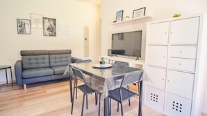 A compact living space with a grey sofa, a dining table with chairs, a flat-screen TV, and a white cabinet with multiple drawers. - Il Giardino Dell Eden- Italian Homing (Sirmione)