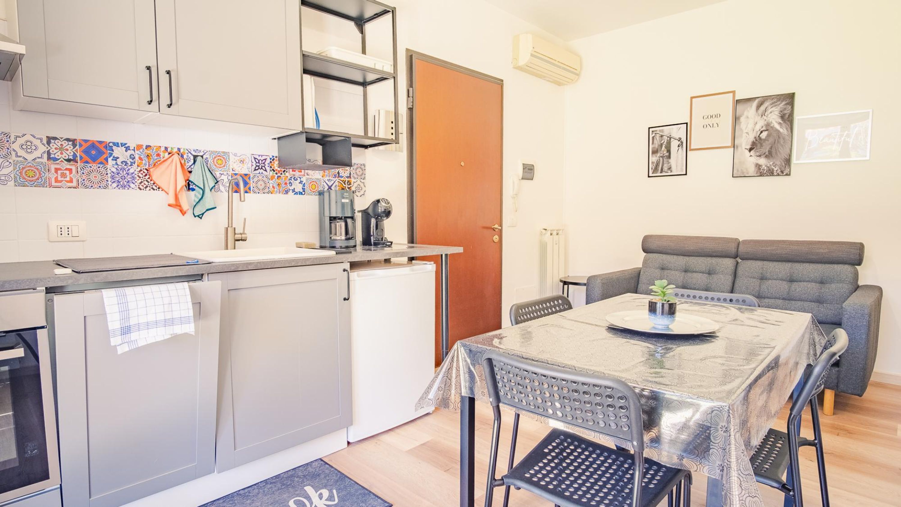 A compact kitchen with a sink, coffee machine, and a small refrigerator. There is a dining table with chairs and a sofa in the adjacent living area.
