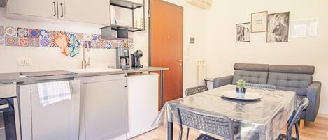 A compact kitchen with a sink, coffee machine, and a small refrigerator. There is a dining table with chairs and a sofa in the adjacent living area.