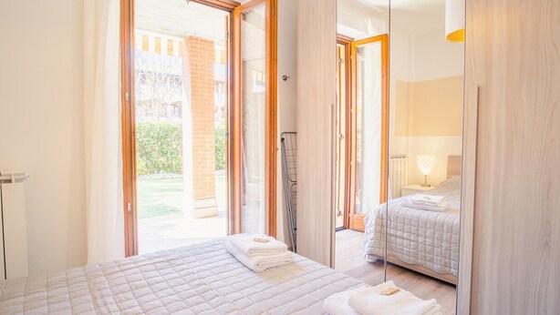 A hotel room with a large bed, a bedside table with a lamp, and a view of a garden through a glass door. - Il Giardino Dell Eden- Italian Homing (Sirmione)