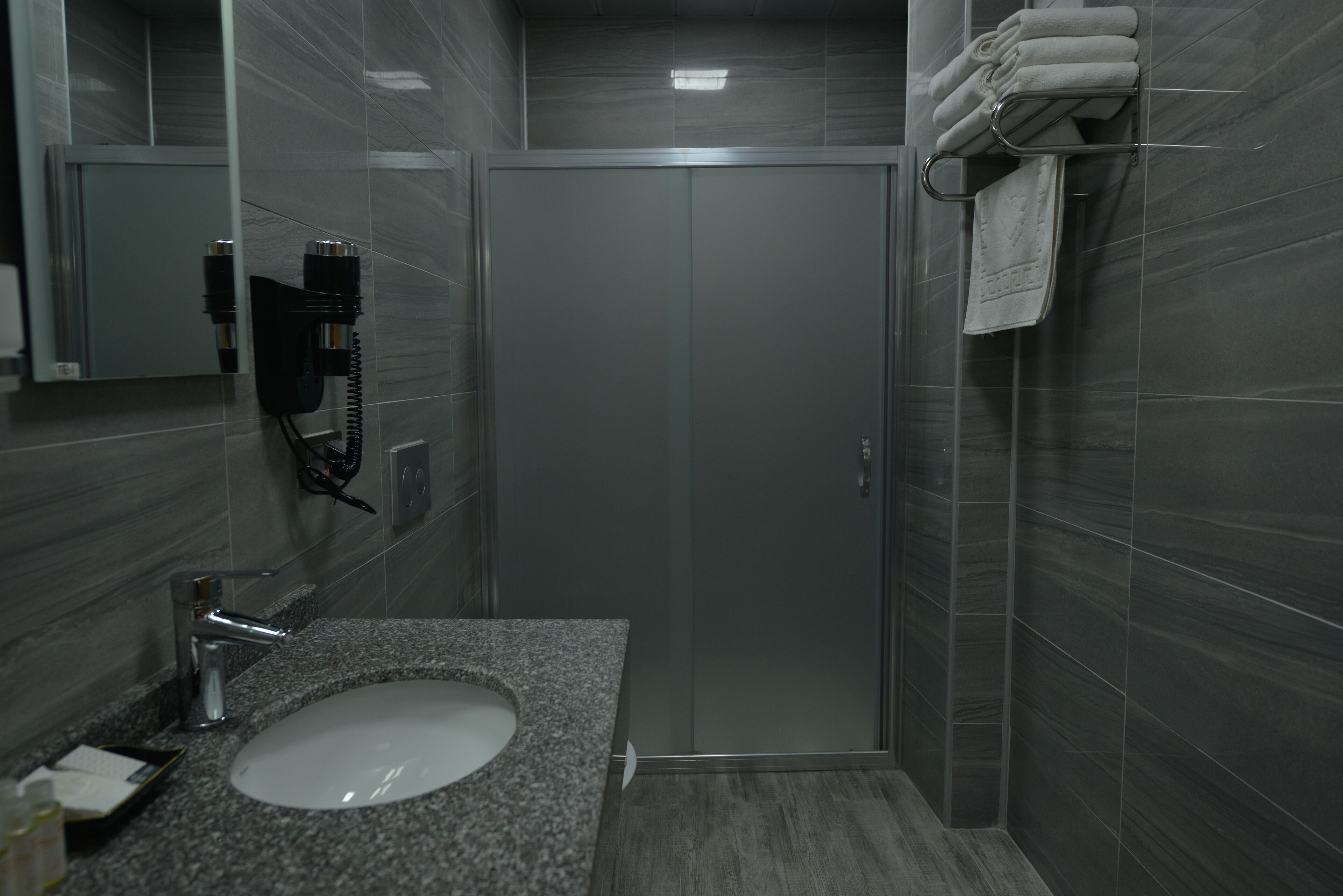 Une salle de bain moderne avec un lavabo doté d’un plan de travail en granit, un sèche-cheveux fixé au mur et un porte-serviettes avec des serviettes pliées.