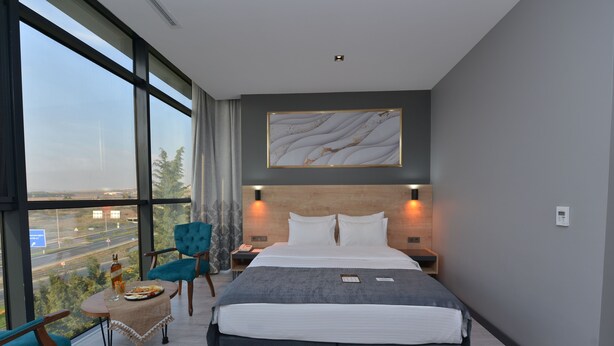 A modern hotel room with a large bed, a view of the cityscape, a small table with a bottle and glasses, and a blue chair. - MILLS HOTEL (Tekirdag)