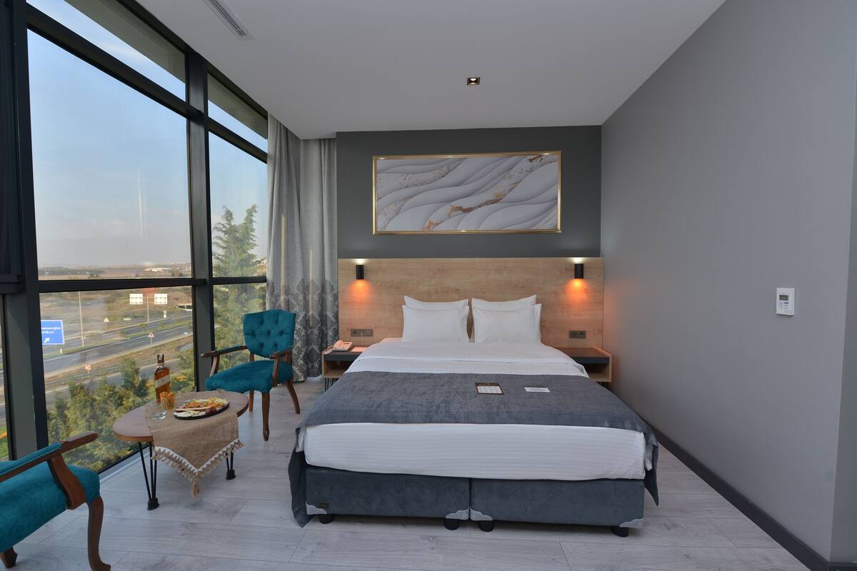 A modern hotel room with a large bed, a view of the cityscape, a small table with a bottle and glasses, and a blue chair.