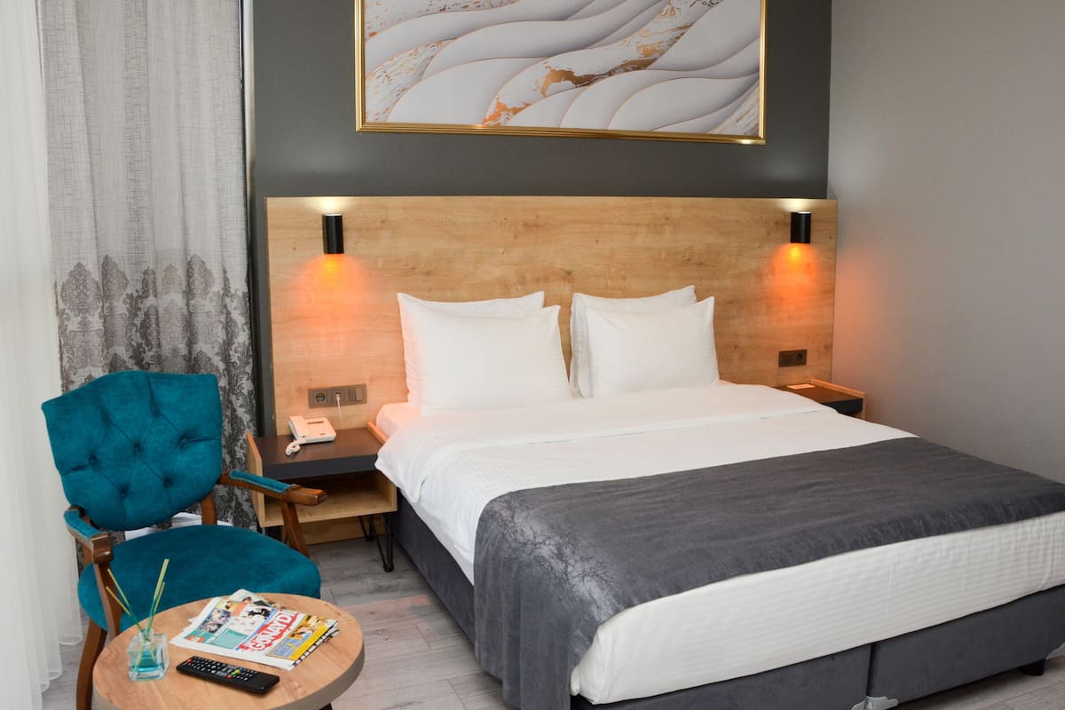 A hotel room with a bed, a nightstand, a chair, a table with a remote control and magazine, and a painting on the wall.