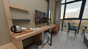 A modern hotel room with a wooden desk, a television, a small refrigerator, a trash bin, and a view of the city through large windows. - MILLS HOTEL (Tekirdag)