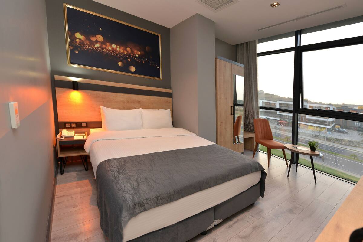 A modern hotel room with a large bed, a nightstand, a desk, a chair, and a large window offering a city view.