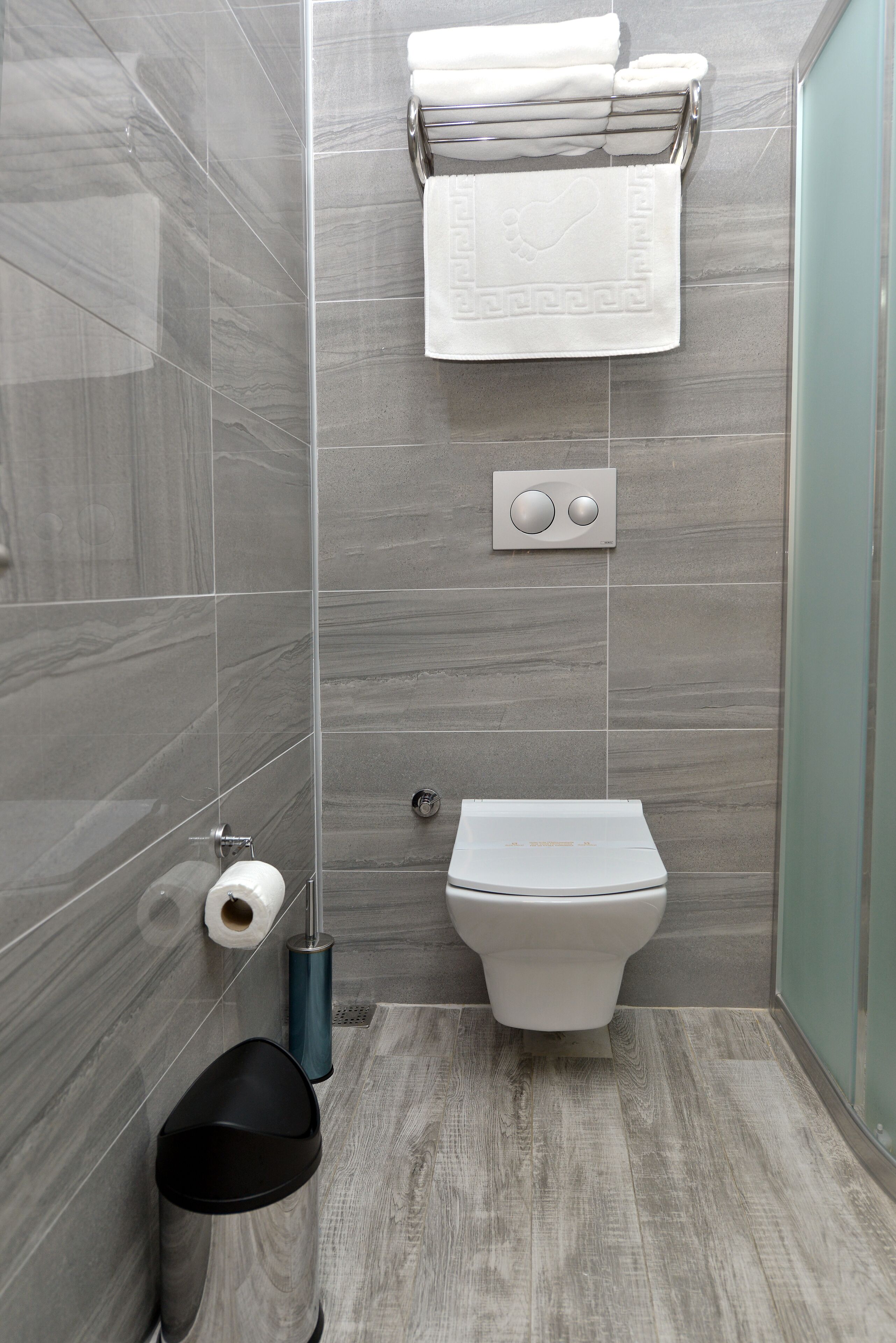 A modern bathroom with grey tiles, a toilet, a trash bin, and a towel rack.