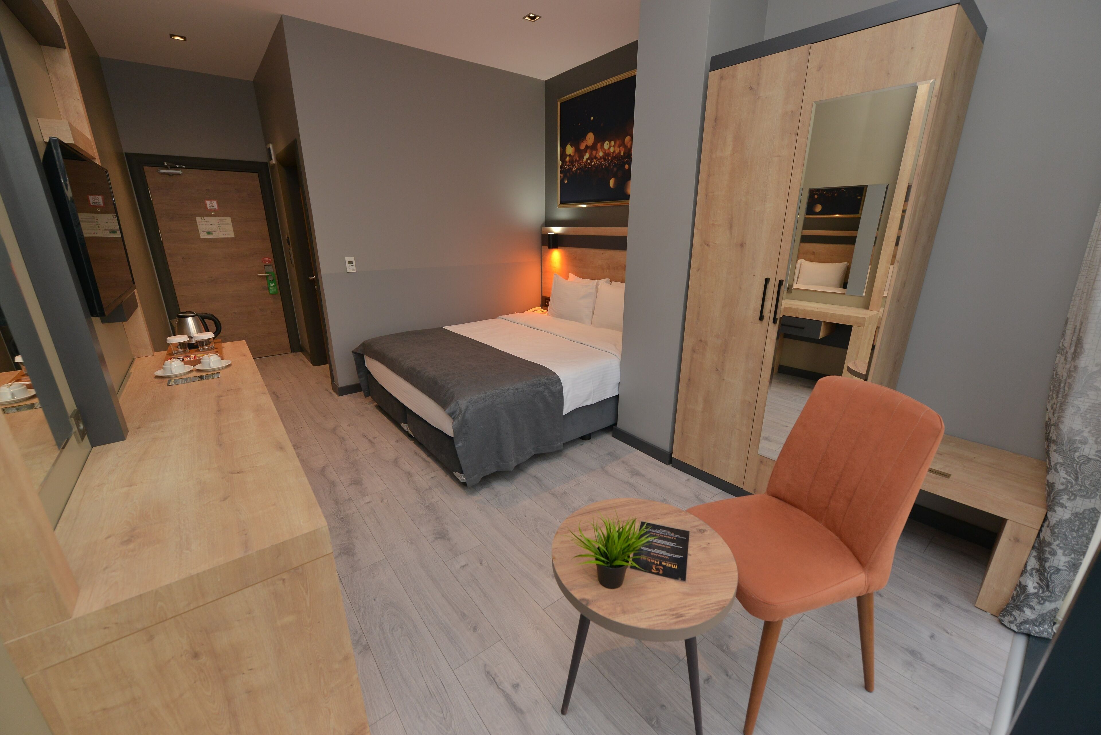 A modern hotel room with a bed, a small table with a plant, a chair, and a wardrobe with a mirror.