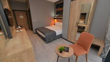 A modern hotel room with a bed, a small table with a plant, a chair, and a wardrobe with a mirror.