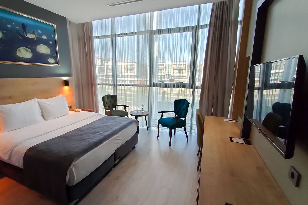 A hotel room with a bed, two chairs, a small table, a TV, and large windows.