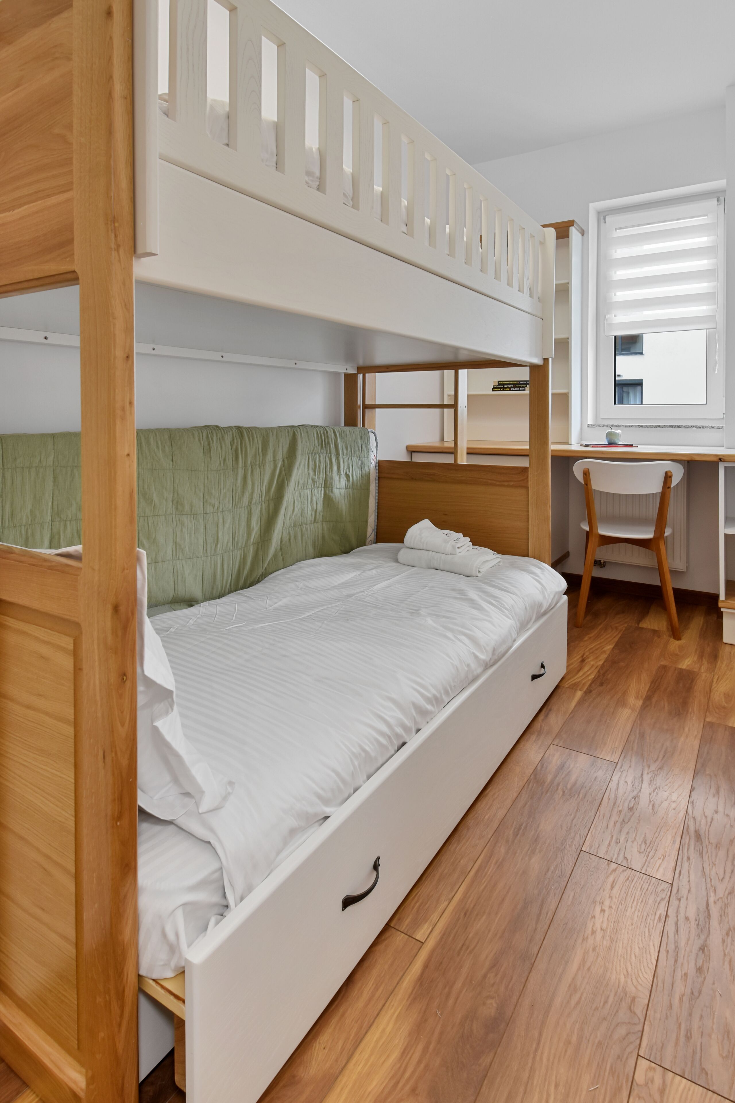 Een stapelbed met een bureau en stoel, houten vloeren en een raam met jaloezieën.