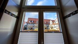 Vista attraverso una finestra con persiane, che mostra un edificio europeo tradizionale con tetto in tegole rosse e diversi piani.