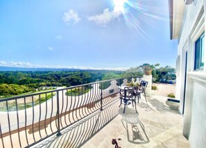 A balcony with a view of a green landscape, a table with chairs, and a railing.