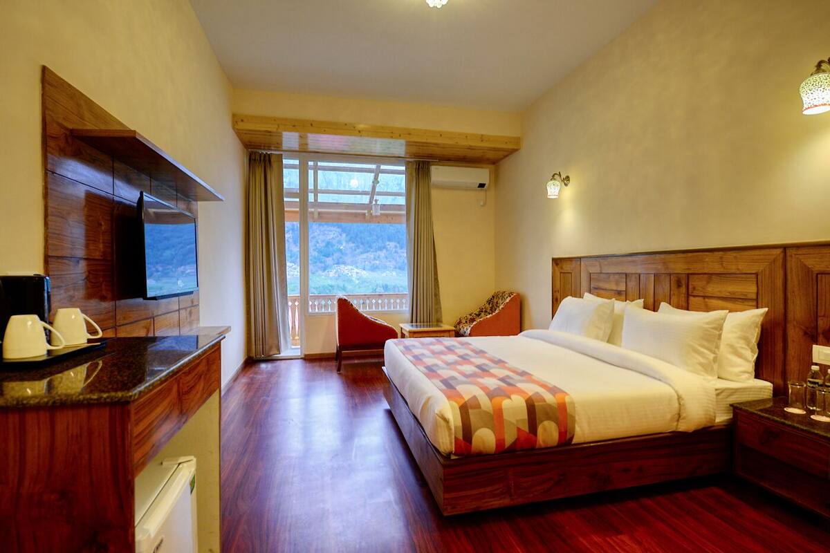 A hotel room with a large bed, a wooden headboard, a television, a desk with a coffee maker, and a balcony with a view of the mountains.