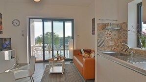 A modern kitchen with a large window, a dining area with a table and chairs, and a living room with a sofa and a TV. - Villa Pettegola in Noto (Noto)