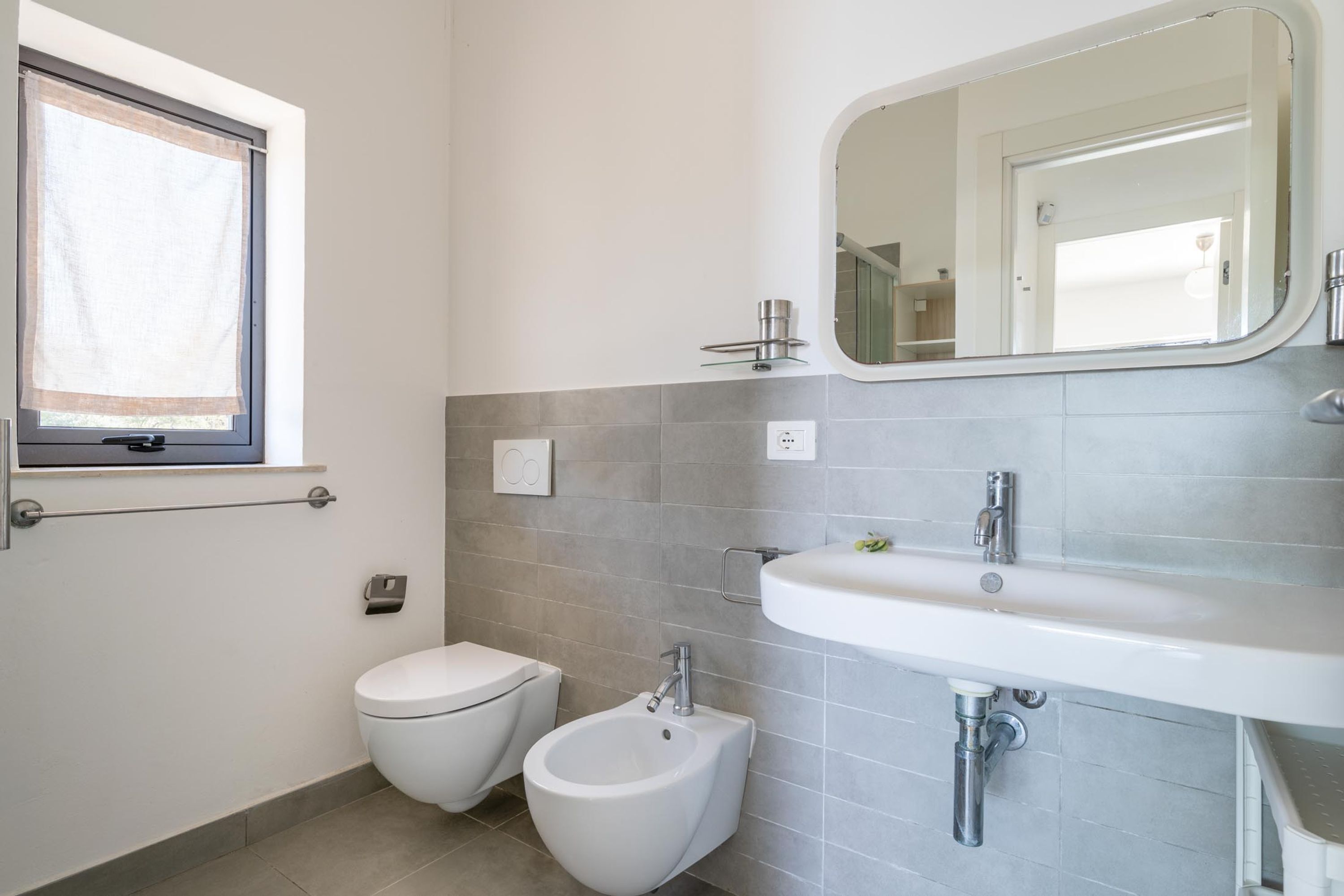A bathroom with a toilet, bidet, sink, mirror, and window.