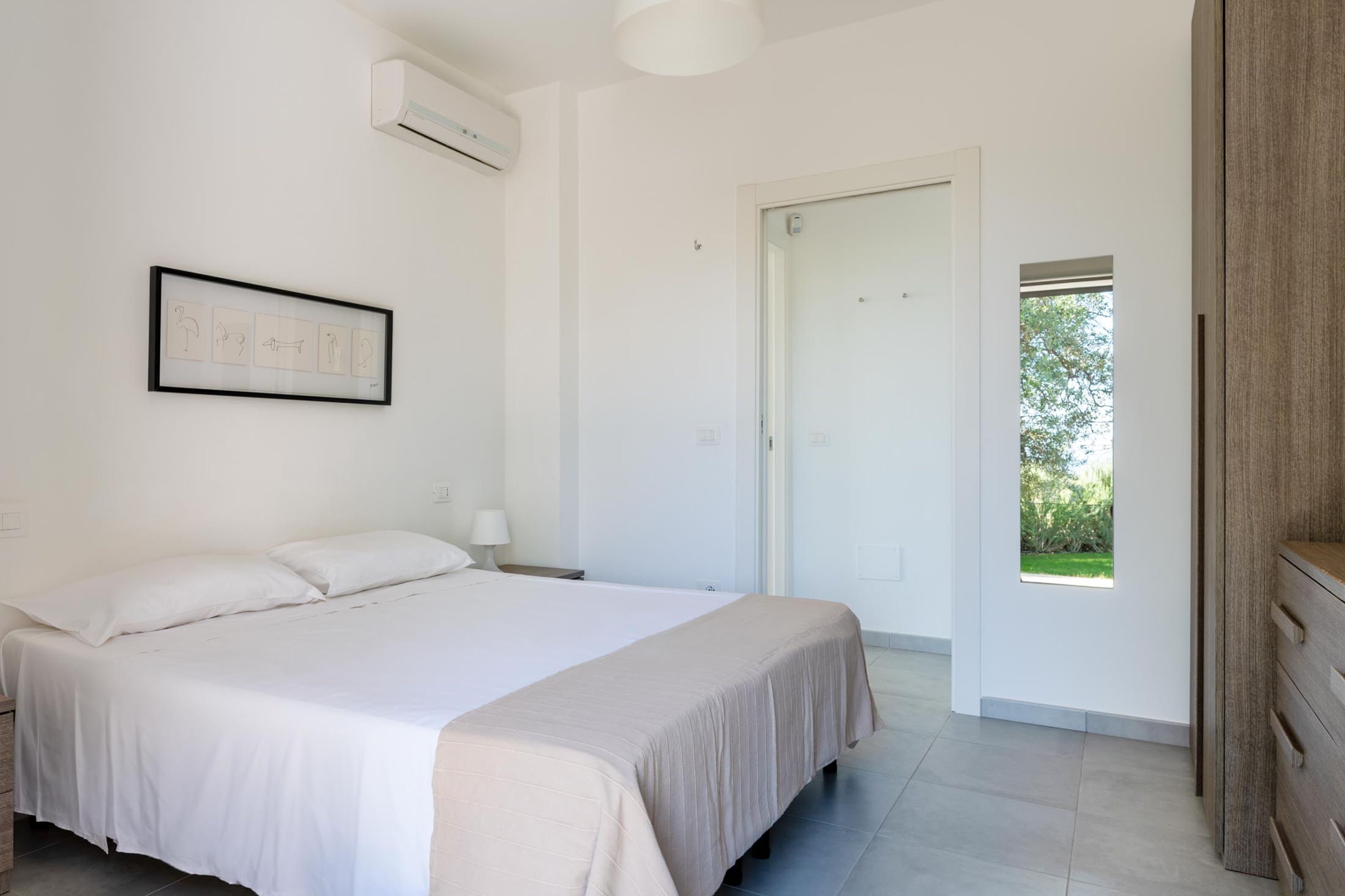 Een slaapkamer met een groot bed, een nachtkastje, een lijst met een afbeelding, een deur naar een balkon en een raam.