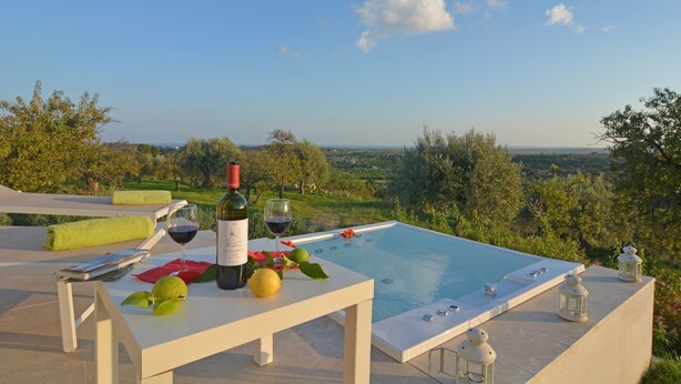 A poolside setting with a table set for a meal, a bottle of wine, and a glass of wine. - Villa Pettegola in Noto (Noto)