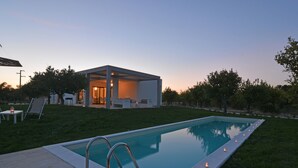 A rectangular swimming pool with a metal ladder, surrounded by a grassy area. There is a small building with a glass door and a patio with chairs and a table. - Villa Papeda in Noto (Noto)